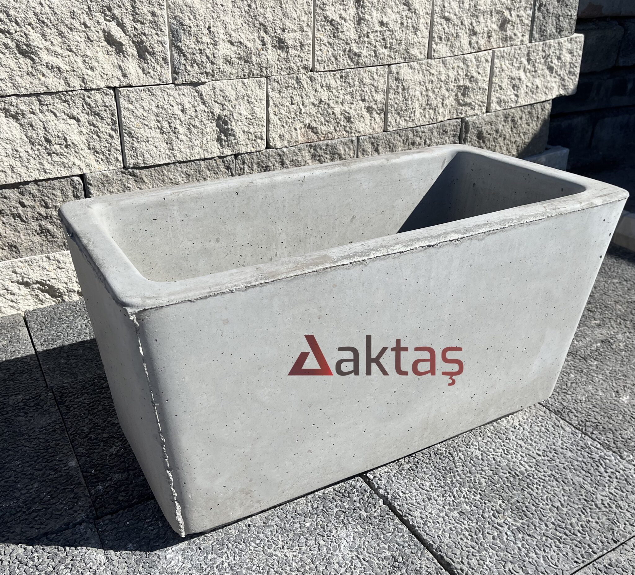 80x40x44 cm boyutlarında, doğal gri renkli mix beton malzemeden yapılmış, dikdörtgen formunda modern bir dış mekan saksısı. Saksının ön yüzünde kırmızı "Aktaş" logosu bulunmaktadır. Saksı boş olup, iç kısmı doğal beton rengindedir. Saksı, koyu gri renkli taş döşeli zemin üzerinde durmakta olup, arka planda açık gri renkli blok taşlardan örülmüş bir duvar görünmektedir. Ürün, dış mekan peyzaj ve güvenlik uygulamaları için uygundur.