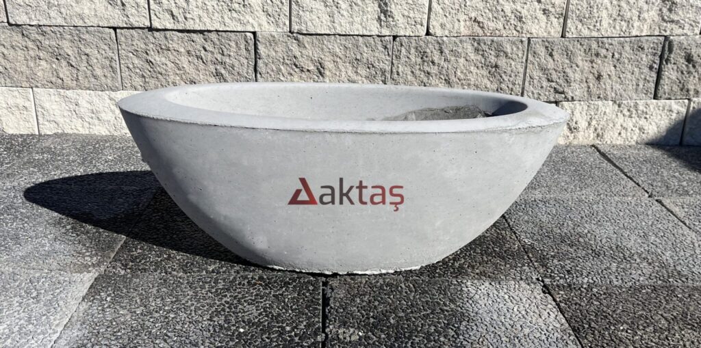80x30x30 cm boyutlarında, doğal beton renginde, yuvarlak hatlara sahip, kayık şeklinde tasarlanmış modern bir beton saksı. Üzerinde Aktaş logosu bulunuyor. Saksı gri taş zemin üzerinde duruyor ve arkasında taş duvar var. Saksı dış mekan kullanımı için uygun.