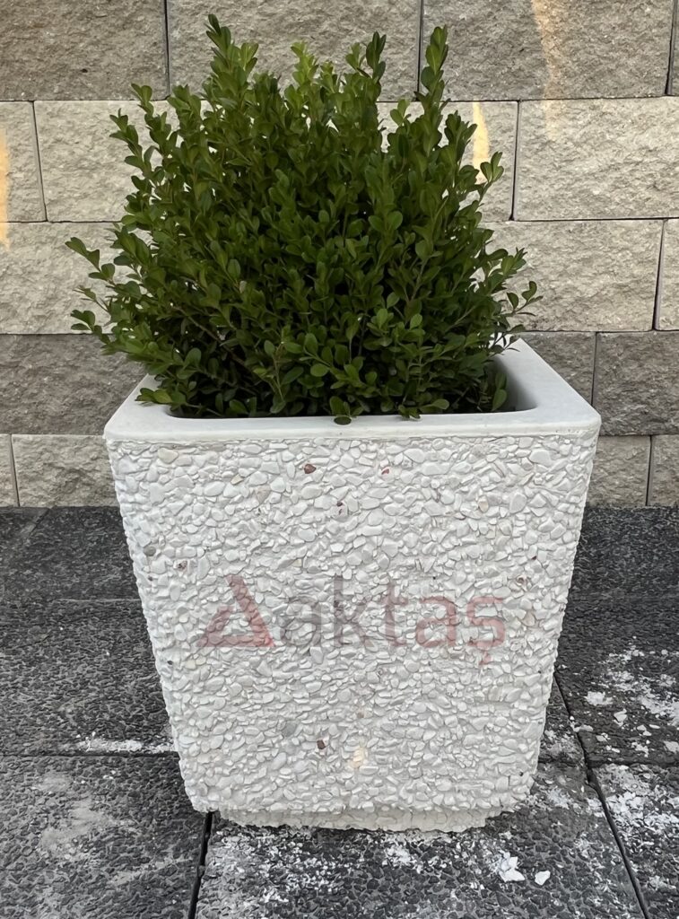 40x40x44 cm boyutlarında, bembeyaz dolomit taş dokulu, kare formunda modern bir dış mekan saksısı. Saksının ön yüzünde kırmızı "Aktaş" logosu bulunmaktadır. Saksının içinde sarı-yeşil yapraklı, gür bir bitki durmaktadır. Saksı, koyu gri renkli taş döşeli zemin üzerinde durmakta olup, arka planda açık renkli tuğla duvardan oluşan bir arka plan görünmektedir. Ürün, dış mekan peyzaj ve güvenlik uygulamaları için uygundur.