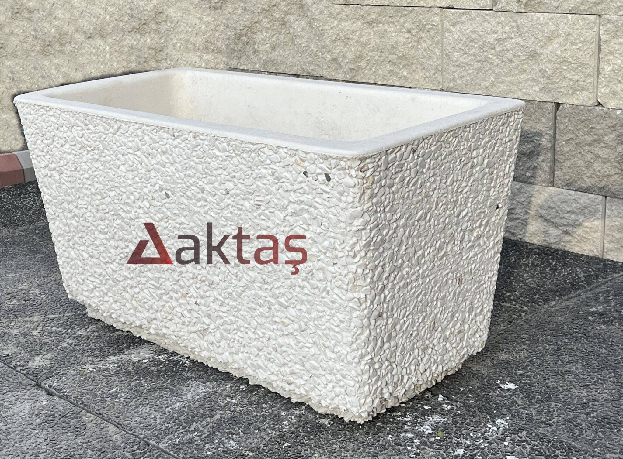 80x40x44 cm boyutlarında, bembeyaz dolomit taş dokulu, dikdörtgen formunda modern bir dış mekan saksısı. Saksının ön yüzünde kırmızı "Aktaş" logosu bulunmaktadır. Saksı boş olup, iç kısmı doğal beton rengindedir. Saksı, koyu gri renkli taş döşeli zemin üzerinde durmakta olup, arka planda açık renkli tuğla duvardan oluşan bir arka plan görünmektedir. Ürün, dış mekan peyzaj ve güvenlik uygulamaları için uygundur.