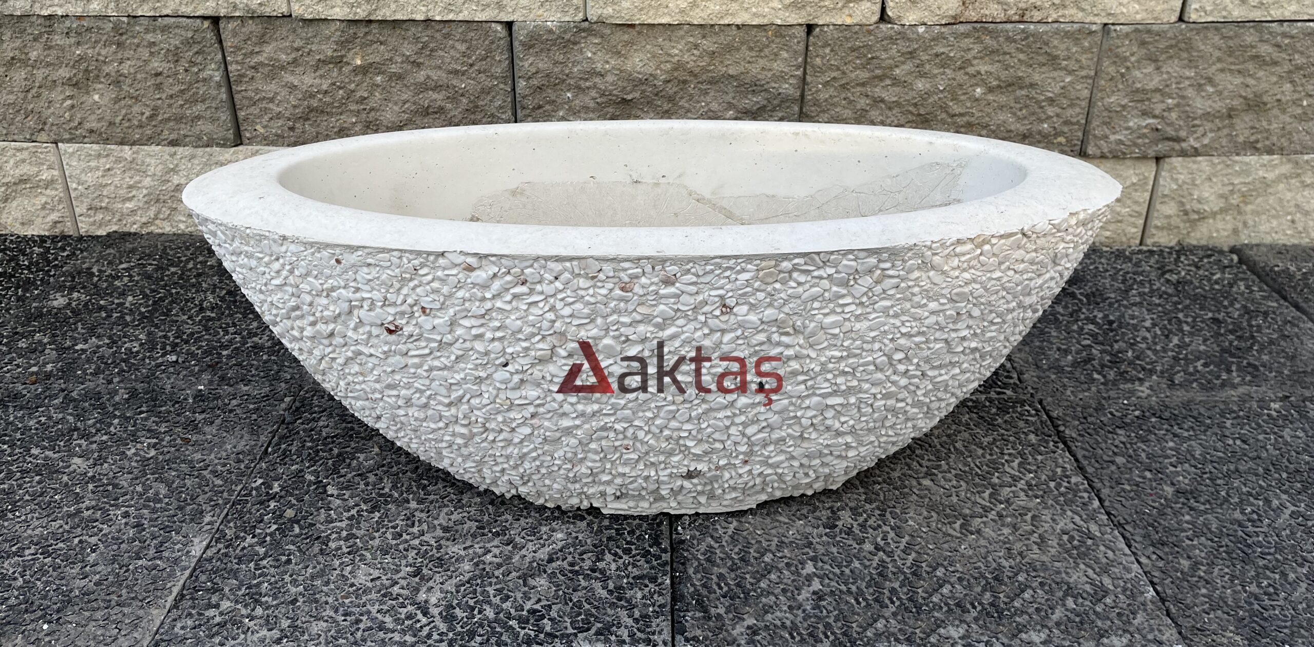 80x30x30 cm boyutlarında, beyaz dolomit taş dokulu, oval ve kayık şeklinde tasarlanmış modern bir dış mekan saksısı. Üzerinde kırmızı Aktaş logosu bulunuyor. Saksı koyu gri taş döşemeli zemin üzerinde duruyor ve arkasında taş duvar var. Saksı, bahçe ve teras kullanımı için uygundur.