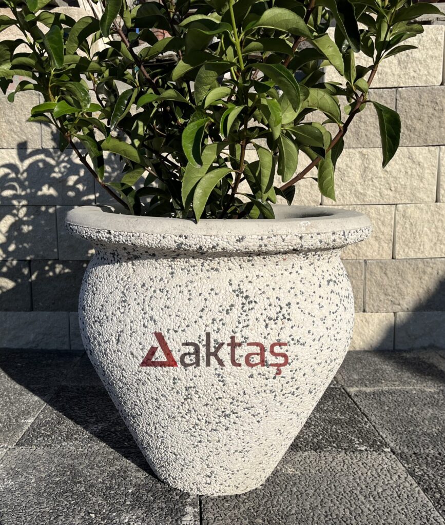 50x50x50 cm boyutlarında, dış yüzeyi beyaz ve gri mozaik parçacıklarıyla kaplı, vazo formunda modern bir dış mekan saksısı. Saksının ön yüzünde kırmızı "Aktaş" logosu bulunmaktadır. Saksının içinde geniş yapraklı, yeşil bir bitki durmaktadır. Saksı, koyu gri renkli taş döşeli zemin üzerinde durmakta olup, arka planda açık gri taşlardan örülmüş bir duvar görünmektedir. Ürün, dış mekan bitkilendirme ve peyzaj için uygundur.