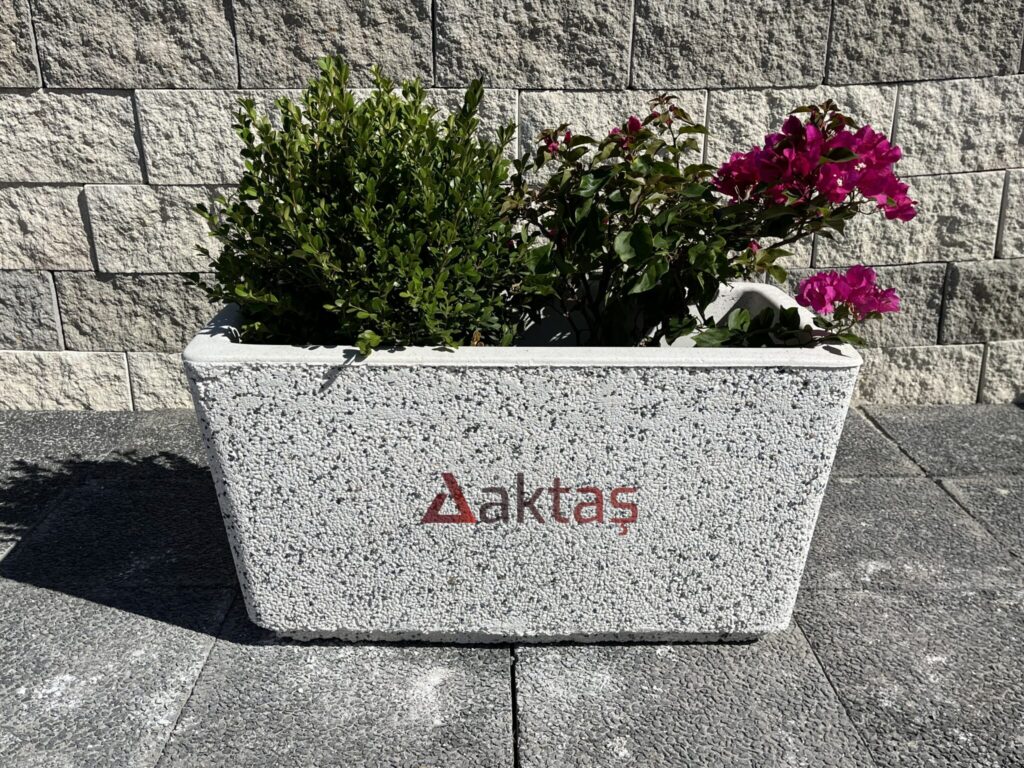 80x40x44 cm boyutlarında, dış yüzeyi beyaz ve gri mozaik parçacıklarıyla kaplı, dikdörtgen formunda modern bir dış mekan saksısı. Saksının ön yüzünde kırmızı "Aktaş" logosu bulunmaktadır. Saksının içinde yeşil yapraklı bir şimşir bitkisi ve pembe çiçekli bir begonvil bitkisi durmaktadır. Saksı, koyu gri renkli taş döşeli zemin üzerinde durmakta olup, arka planda açık gri renkli blok taşlardan örülmüş bir duvar görünmektedir. Ürün, dış mekan peyzaj ve güvenlik uygulamaları için uygundur.