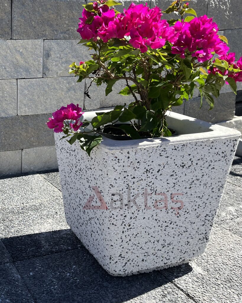 40x40x44 cm boyutlarında, dış yüzeyi beyaz ve gri mozaik parçacıklarıyla kaplı, kare formunda modern bir dış mekan saksısı. Saksının ön yüzünde kırmızı "Aktaş" logosu bulunmaktadır. Saksının içinde pembe çiçekli, gür yeşil yapraklı bir bitki durmaktadır. Saksı, koyu gri renkli taş döşeli zemin üzerinde durmakta olup, arka planda açık renkli tuğla duvardan oluşan bir arka plan görünmektedir. Ürün, dış mekan peyzaj ve güvenlik uygulamaları için uygundur.
