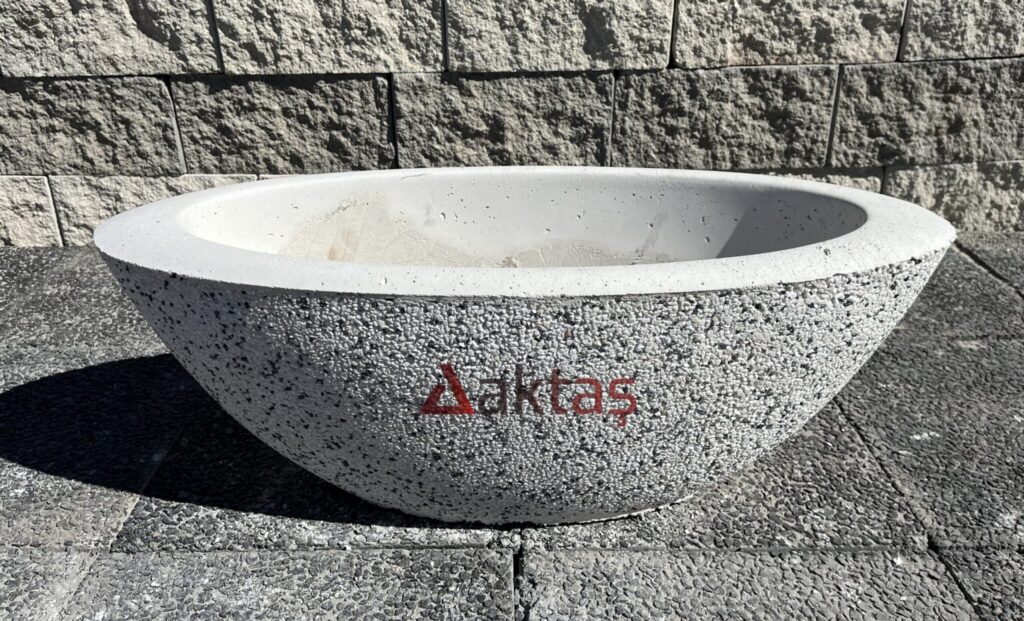 80x30x30 cm boyutlarında, beyaz küçük mozaik parçacıklarıyla kaplı, yuvarlak hatlara sahip, kayık şeklinde modern bir beton saksı. Üzerinde kırmızı Aktaş logosu bulunuyor. Saksı koyu gri taş döşemeli zemin üzerinde duruyor ve arkasında taş duvar var. Saksı, dış mekan ve bahçe dekorasyonu için uygundur.