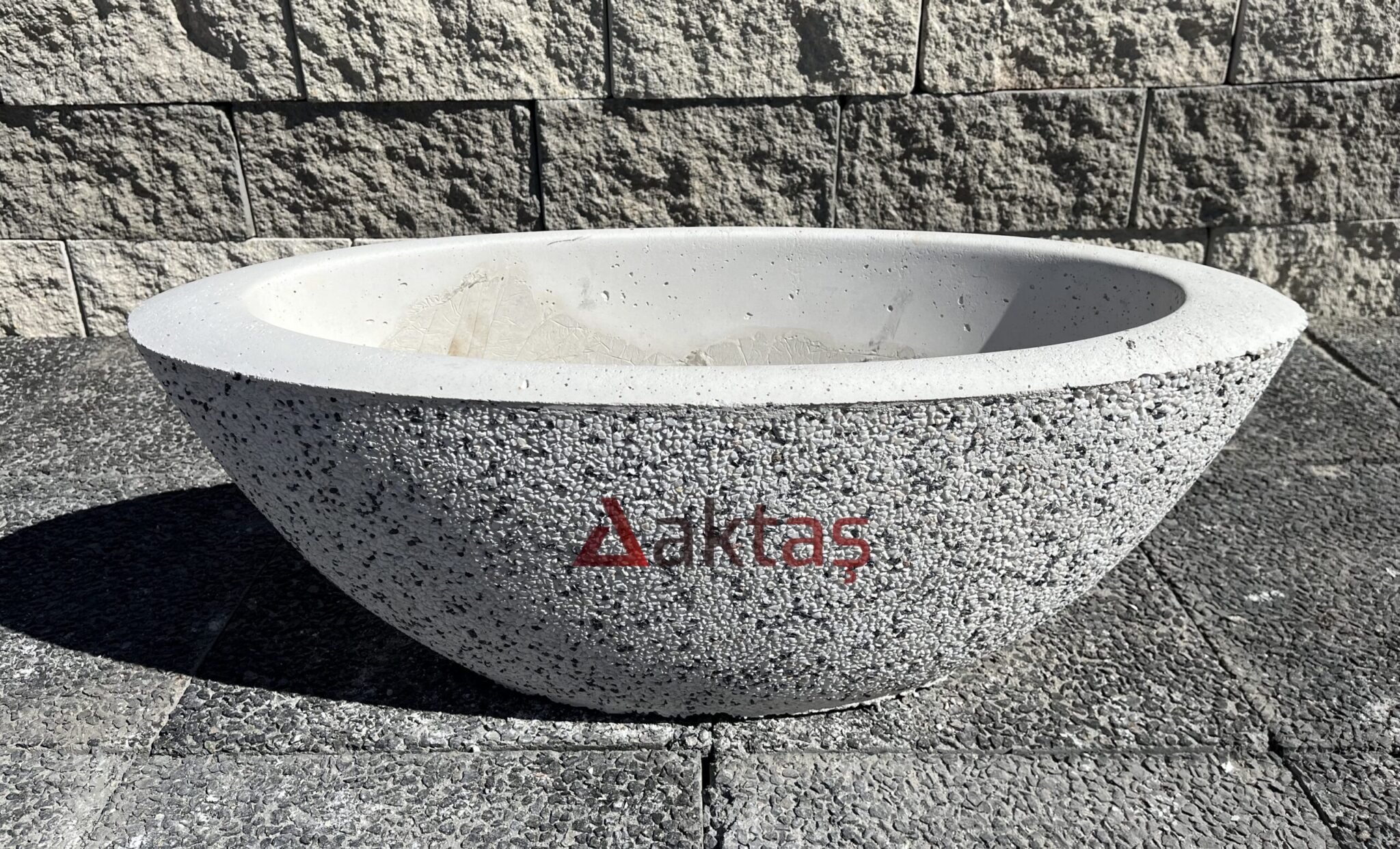 80x30x30 cm boyutlarında, beyaz küçük mozaik parçacıklarıyla kaplı, yuvarlak hatlara sahip, kayık şeklinde modern bir beton saksı. Üzerinde kırmızı Aktaş logosu bulunuyor. Saksı koyu gri taş döşemeli zemin üzerinde duruyor ve arkasında taş duvar var. Saksı, dış mekan ve bahçe dekorasyonu için uygundur.