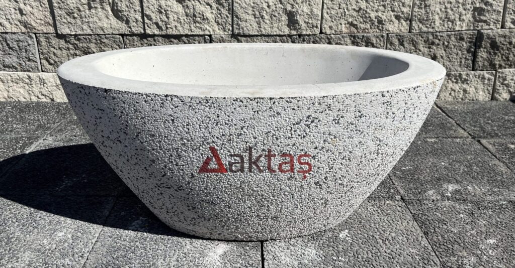 90x45x40 cm boyutlarında, dış yüzeyi beyaz ve gri mozaik parçacıklarıyla kaplı, oval ve kayık formunda, büyük bir beton saksı. Saksının ön yüzünde kırmızı Aktaş logosu yazısı bulunmaktadır. Saksı, koyu renkli taş döşemeli bir zemin üzerinde durmakta olup, arka planda gri taşlardan örülmüş bir duvar görünmektedir. Ürün, geniş dış mekan peyzaj düzenlemeleri için uygundur.