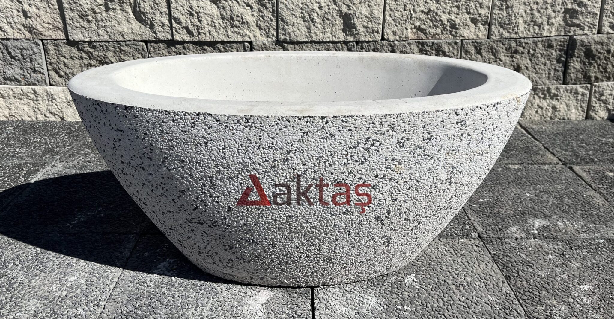 90x45x40 cm boyutlarında, dış yüzeyi beyaz ve gri mozaik parçacıklarıyla kaplı, oval ve kayık formunda, büyük bir beton saksı. Saksının ön yüzünde kırmızı Aktaş logosu yazısı bulunmaktadır. Saksı, koyu renkli taş döşemeli bir zemin üzerinde durmakta olup, arka planda gri taşlardan örülmüş bir duvar görünmektedir. Ürün, geniş dış mekan peyzaj düzenlemeleri için uygundur.