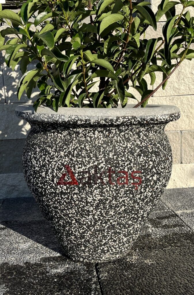 50x50x50 cm boyutlarında, dış yüzeyi siyah ve beyaz mozaik parçacıklarıyla kaplı, vazo formunda modern bir dış mekan saksısı. Saksının ön yüzünde kırmızı "Aktaş" logosu bulunmaktadır. Saksının içinde gür yeşil yapraklı bir bitki durmaktadır. Saksı, koyu renkli taş döşeli zemin üzerinde durmakta olup, arka planda açık renkli tuğla duvardan oluşan bir arka plan görünmektedir. Ürün, dış mekan bitkilendirme ve peyzaj için uygundur.