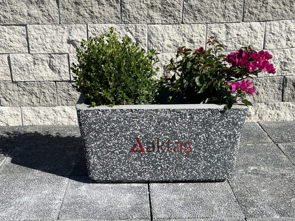 80x40x44 cm boyutlarında, dış yüzeyi siyah ve beyaz mozaik parçacıklarıyla kaplı, dikdörtgen formunda modern bir dış mekan saksısı. Saksının ön yüzünde kırmızı "Aktaş" logosu bulunmaktadır. Saksının içinde yeşil yapraklı bir şimşir bitkisi ve pembe çiçekli bir begonvil bitkisi durmaktadır. Saksı, açık renkli taş duvar önünde, koyu gri renkli kaldırım taşları üzerinde durmaktadır. Ürün, dış mekan peyzaj ve güvenlik uygulamaları için uygundur.