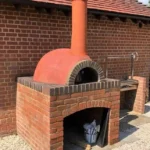 Kırmızı tuğla kaide ve kiremit rengi sıva kubbesi olan, barbekü eklentili, modern dış mekan taş fırını.