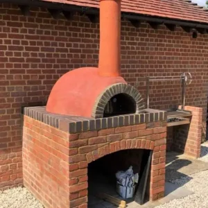 Kırmızı tuğla kaide ve kiremit rengi sıva kubbesi olan, barbekü eklentili, modern dış mekan taş fırını.
