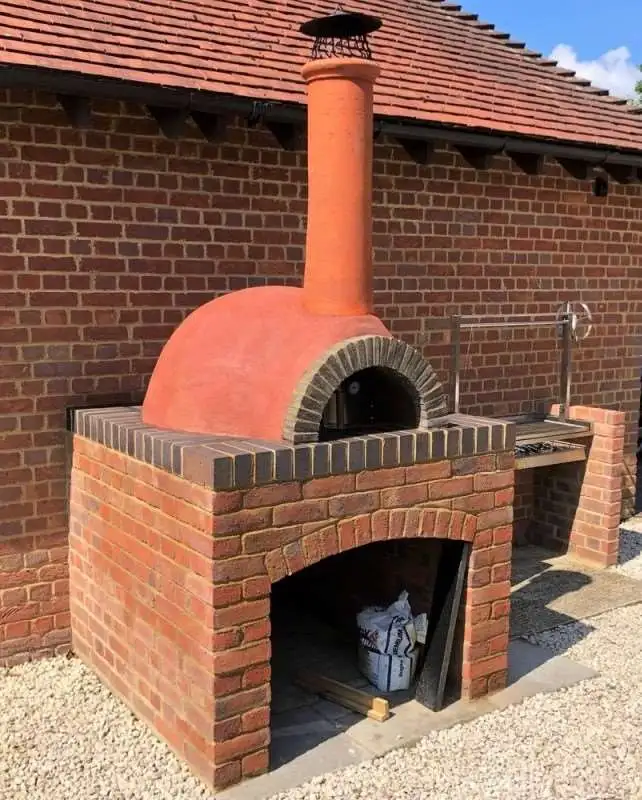 Kırmızı tuğla kaide ve kiremit rengi sıva kubbesi olan, barbekü eklentili, modern dış mekan taş fırını.
