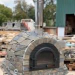 Doğal kayrak taşlarıyla kaplanmış, siyah metal kapaklı ve paslanmaz çelik bacalı hazır taş fırın (pizza fırını).