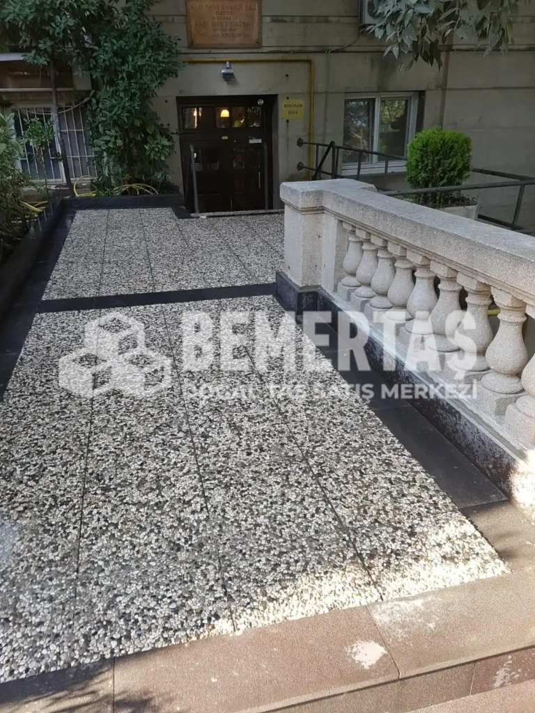 Siyah bordür ile çerçevelenmiş, bej ve gri çakıllı 40x40 wash beton karo ile kaplanmış bina giriş rampası ve avlusu.