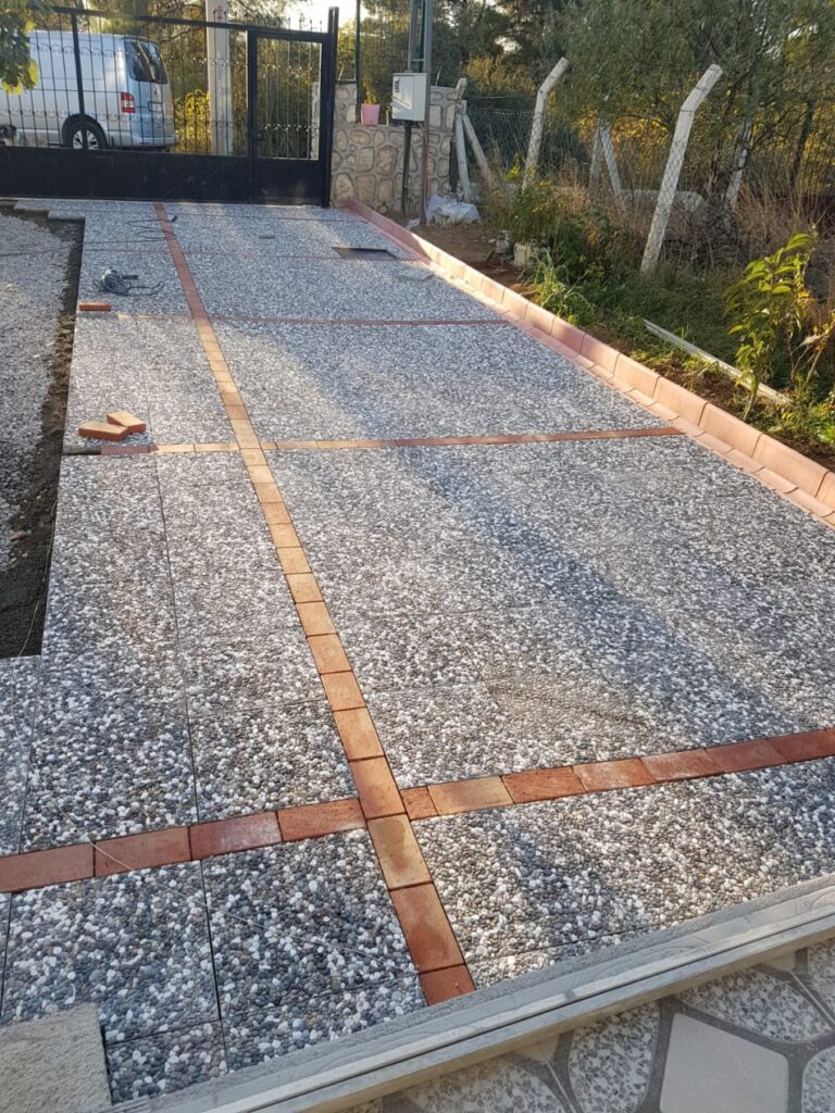 Kırmızı tuğla bordürlerle çapraz kesilmiş ve döşenmiş doğal çakıl agregalı wash beton karodan yapılmış araç giriş yolu.